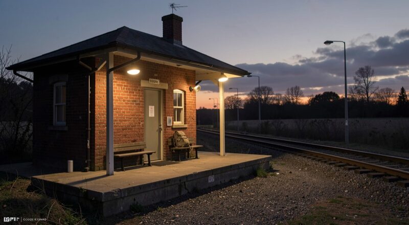 A small unstaffed brick rail depot at dusk, its platform lit by two sodium lamps, a single backpack on a bench, tracks leading away